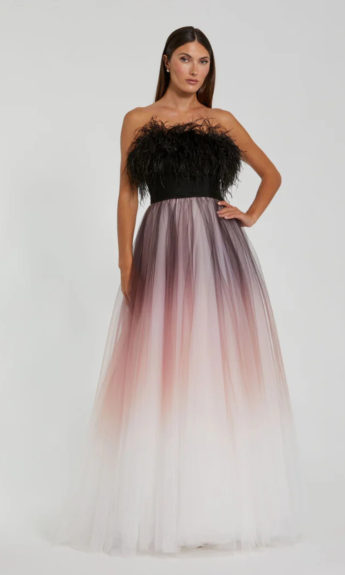 A woman wears the Long Formal Dress 20557 by Mac Duggal, featuring a strapless black feathered bodice and ombre feather-trimmed floor-length tulle skirt that fades from black to pink to white. She stands against a plain background.